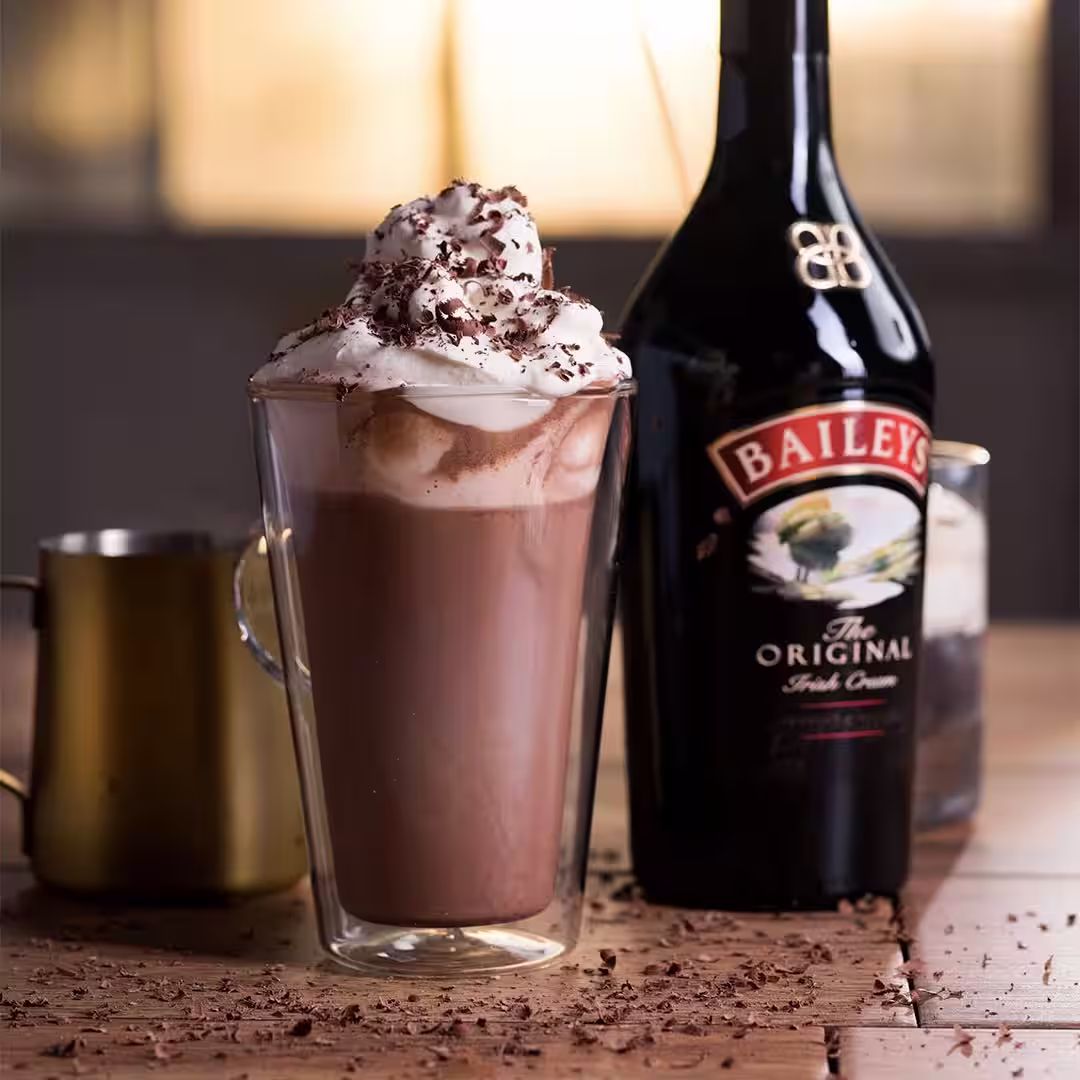 Er is een Baileys warme chocolademelk in een hoog glas, naast een Baileys fles. Hij is zacht en overgoten met slagroom en chocolade.
