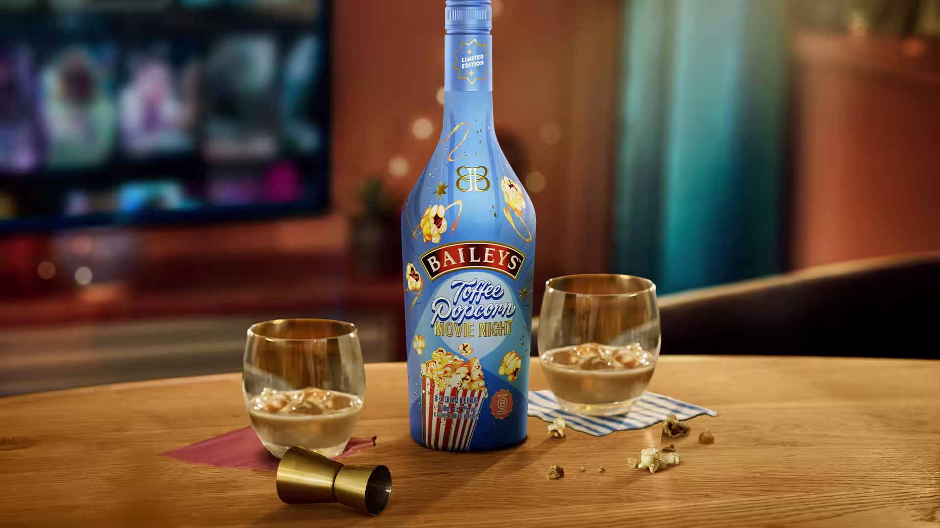 Baileys Toffee Popcorn bottle with two caramel-drizzled ice cream scoops topped with popcorn. The background is cosy with a home movie night setting.