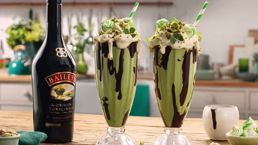 Two tall Baileys St Patrick’s Day Shake with chocolate drizzle, whipped cream and green straws, with a Baileys bottle in the background.