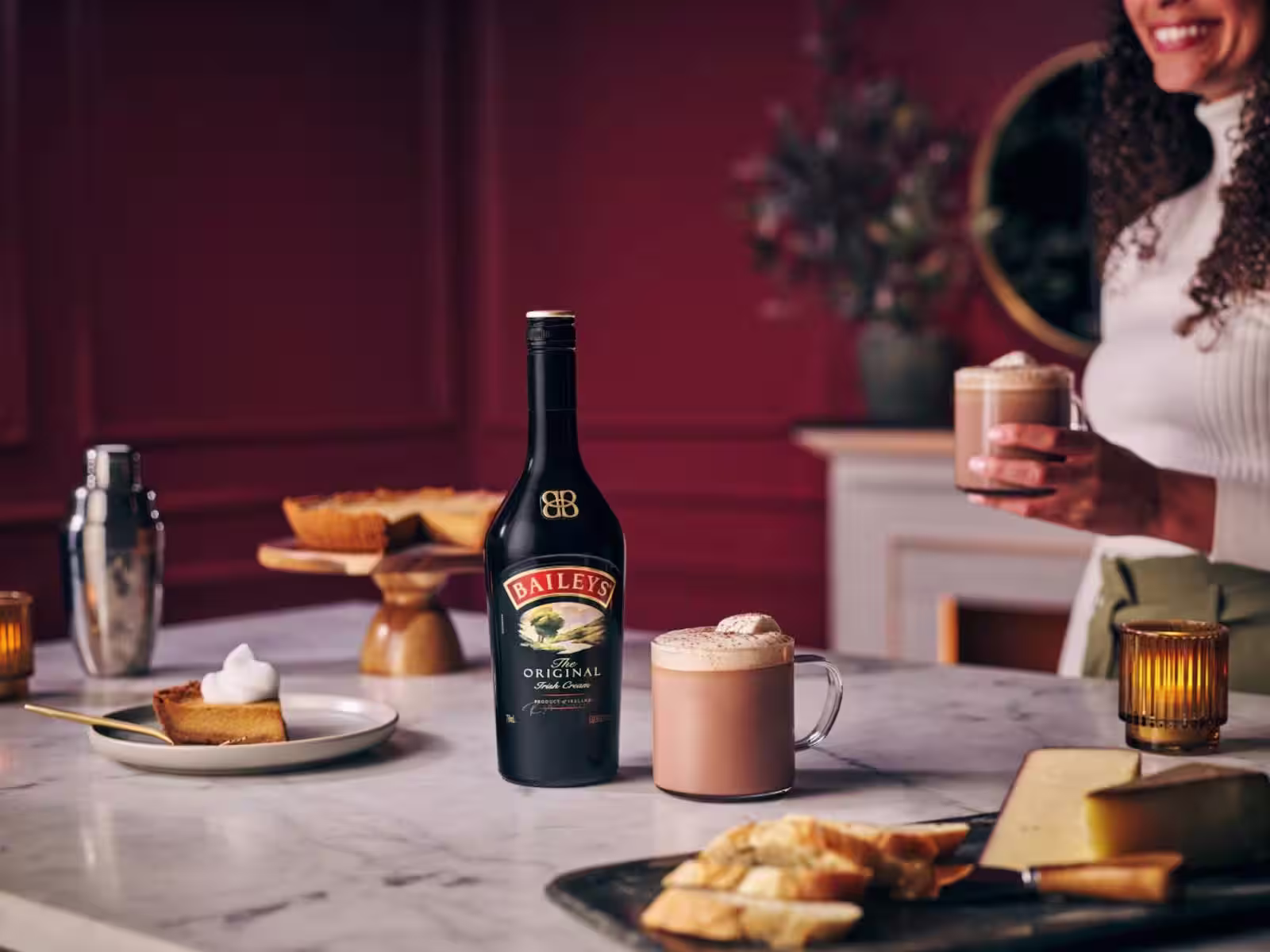 A Baileys hot chocolate sits on a marble table, surrounded by sweet treats and is next to a Baileys bottle. A smiling woman holds her tasty hot choc.