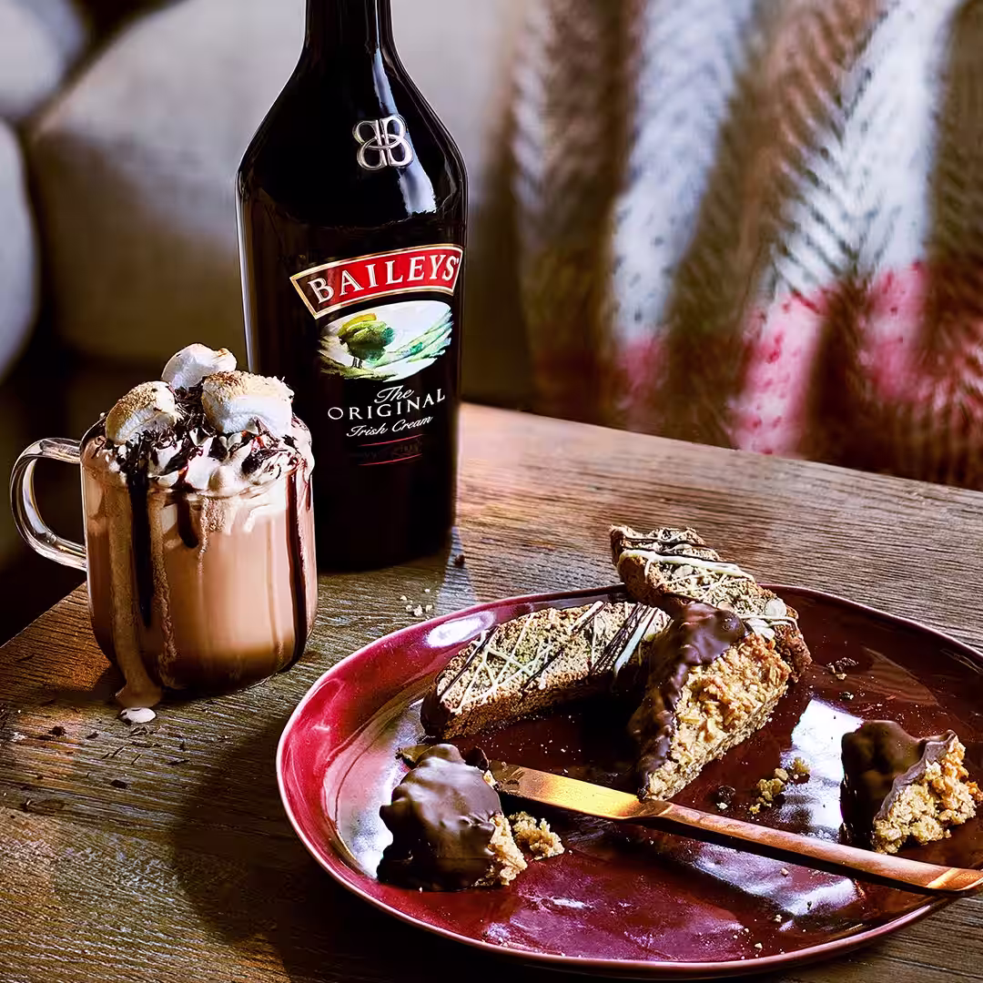 A Baileys hot chocolate sits on a table in a cosy living room. It's topped with marshmallows, cream and chocolate drips.