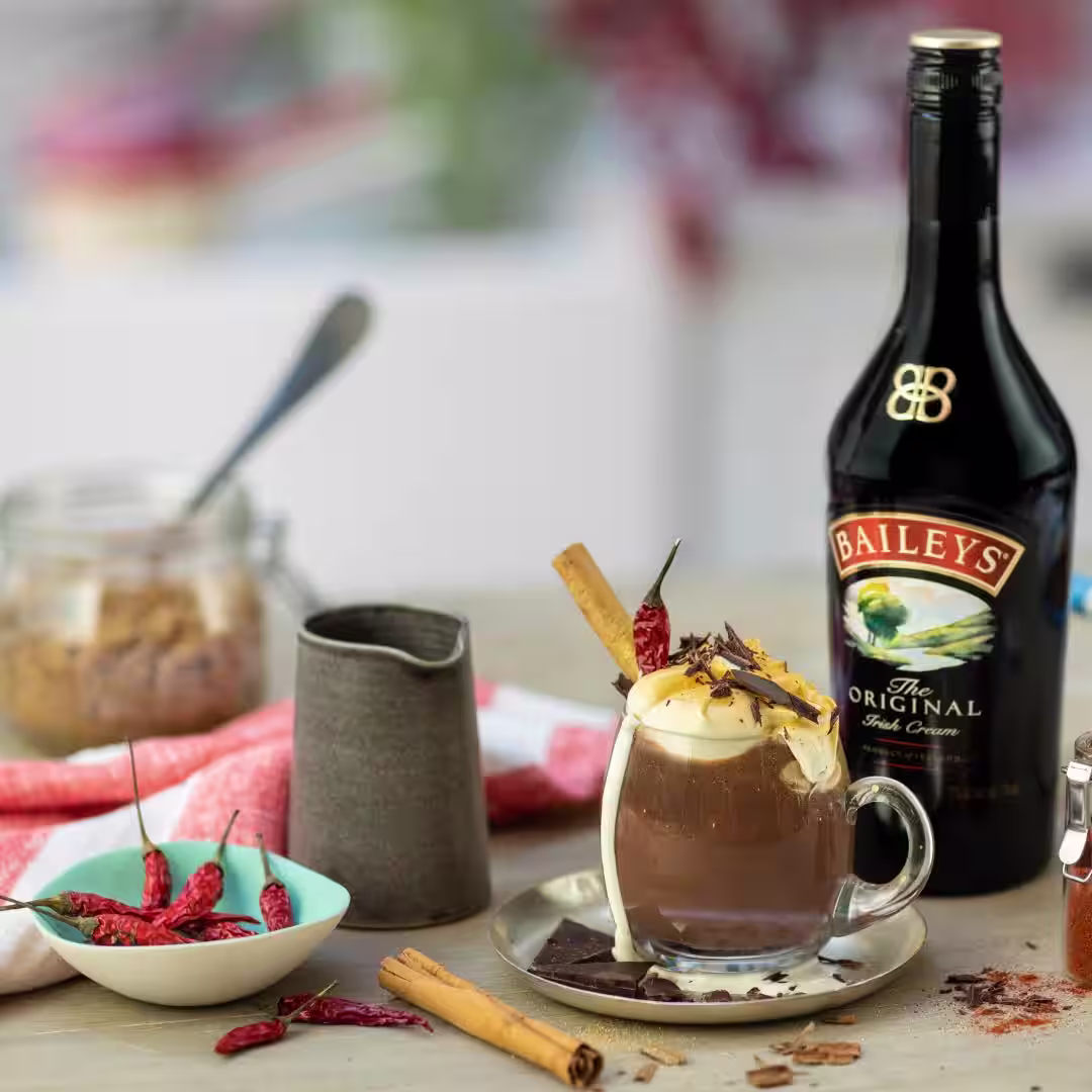A sweet n' spicy Baileys hot chocolate sits on a counter. Surrounded by Baileys, cinnamon sticks and chillies - yummy!